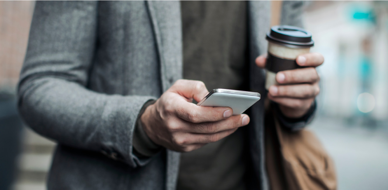 A young man uses his cellphone while he walks down the street with a coffee in hand.