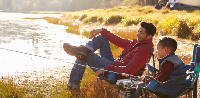 On the edge of a lake in the fall, a happy father sits with his little boy who is holding a large fishing rod. 