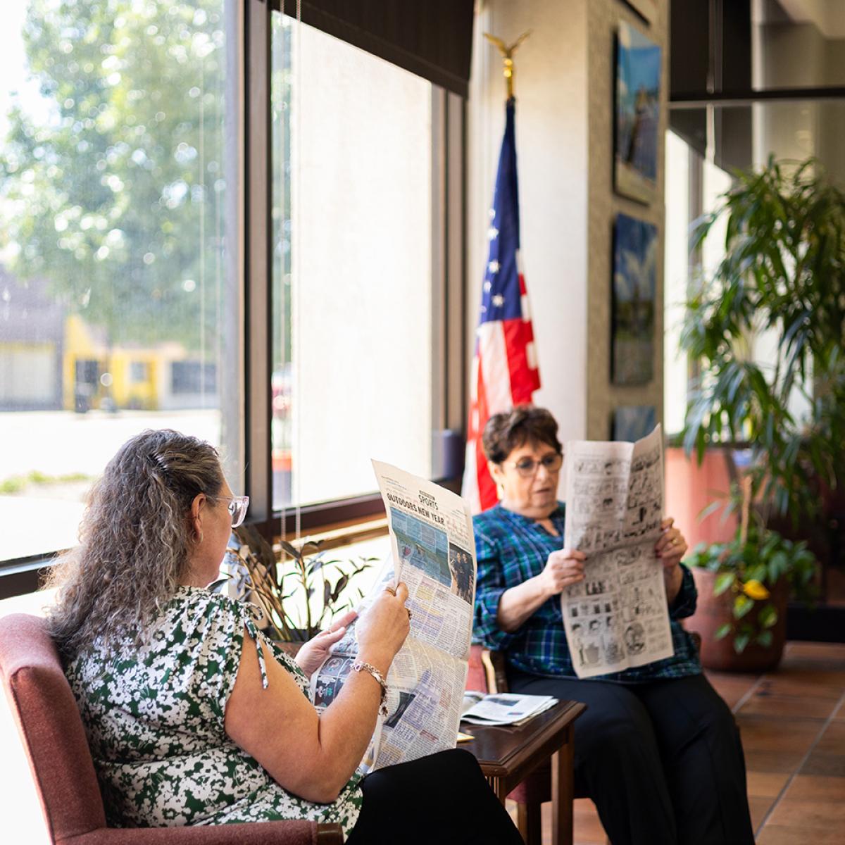 Two women sitting in chairs reading newspapers by the window